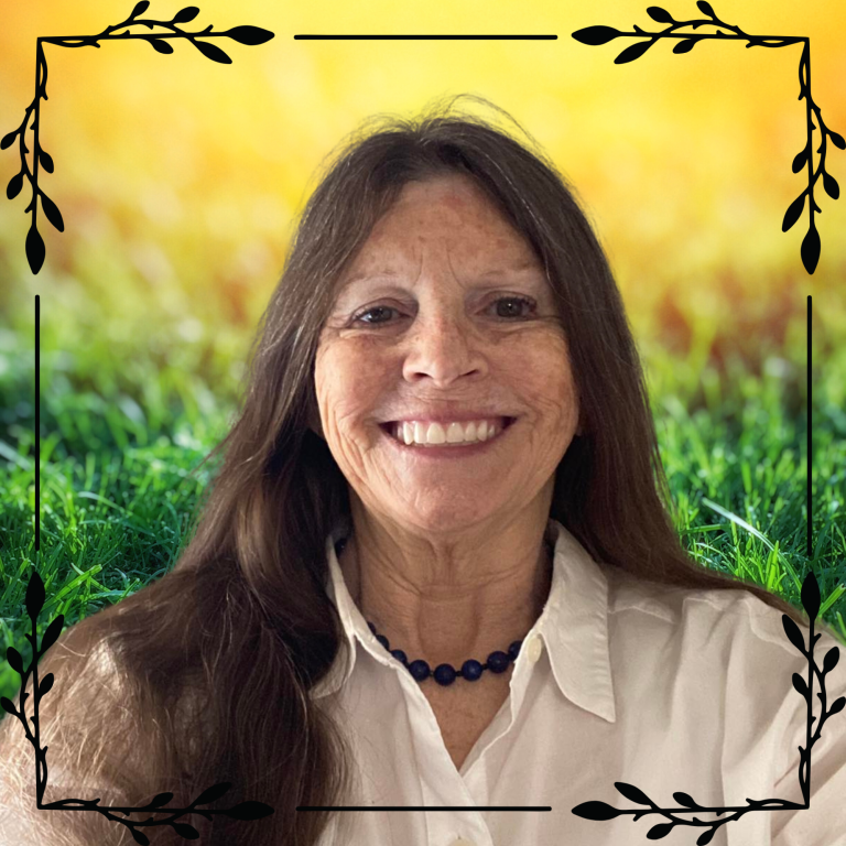 Founder of TriFourOne, Sheila Shafer Smiling woman with long hair, wearing a necklace, in front of a blurred outdoor background.